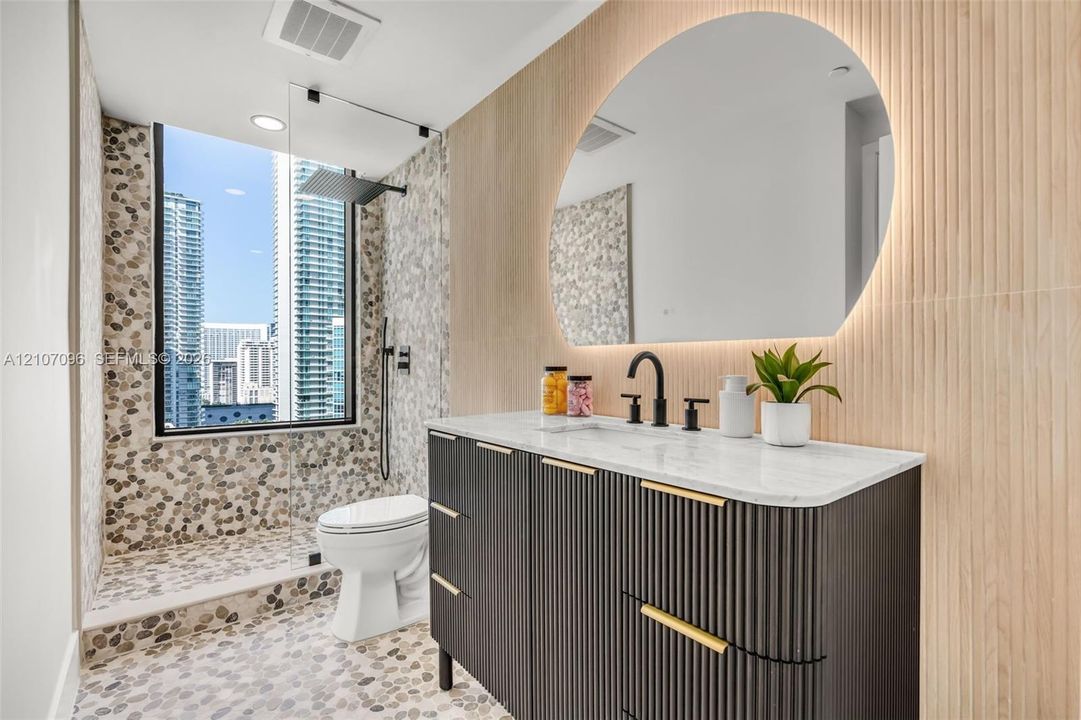 Custom dark fluted vanity with gold-tone hardware and light-toned stone countertop, integrated backlit arched mirror, floor-to-ceiling pebble tile shower surround, and matte black rain shower head