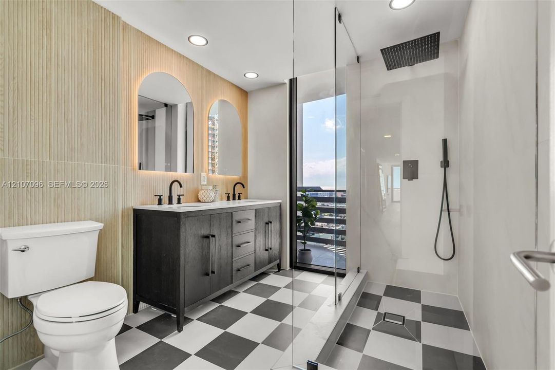 Bathroom featuring a dark wood-finish vanity with dual undermount sinks, backlit arched mirrors, checkerboard tile flooring, and a walk-in shower with a matte black rainfall showerhead