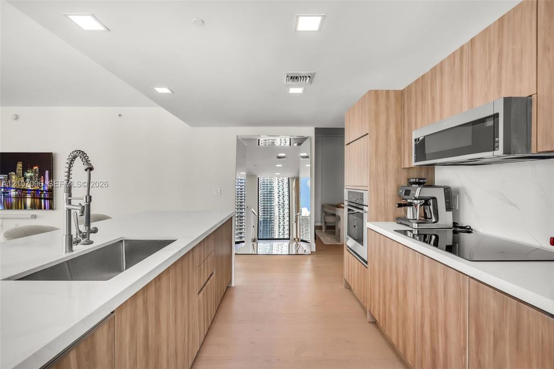 Kitchen featuring wood-finish flat-panel cabinetry, light-toned stone-finish countertops, stainless steel undermount sink with high-arc faucet, integrated wall oven, and built-in microwave
