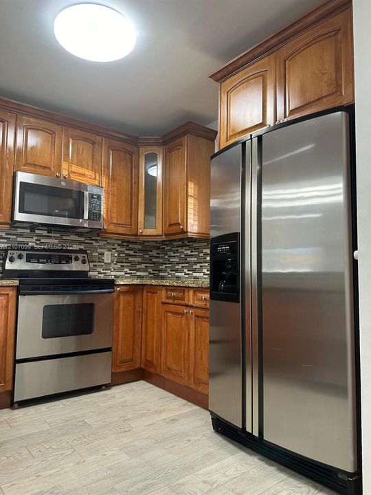 Kitchen featuring wood cabinetry, stainless steel appliances, stone-look countertops, a mosaic tile backsplash, and wood-finish flooring
