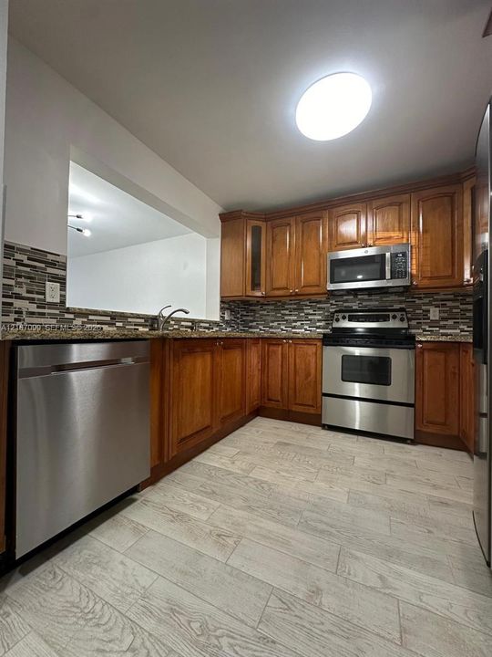 Kitchen featuring wood cabinetry, stone countertops, multi-toned tile backsplash, stainless steel appliances, and wood-finish tile flooring