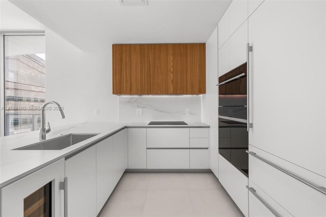 Modern kitchen featuring flat-panel white cabinetry, wood-finish upper cabinets, stone-surfaced countertops with matching backsplash, integrated wall oven, and stainless steel under-mount sink