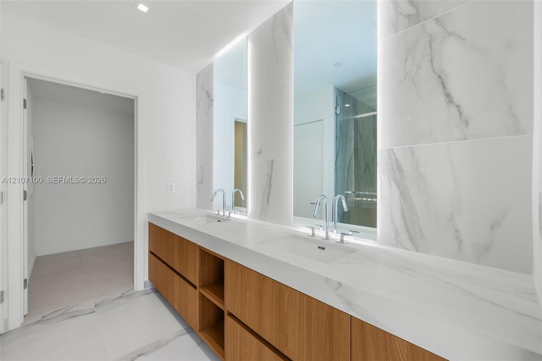 Dual-sink vanity featuring a light-toned countertop, wood-finish cabinetry, floor-to-ceiling veined tile wall treatment, and integrated LED-backlit mirrors