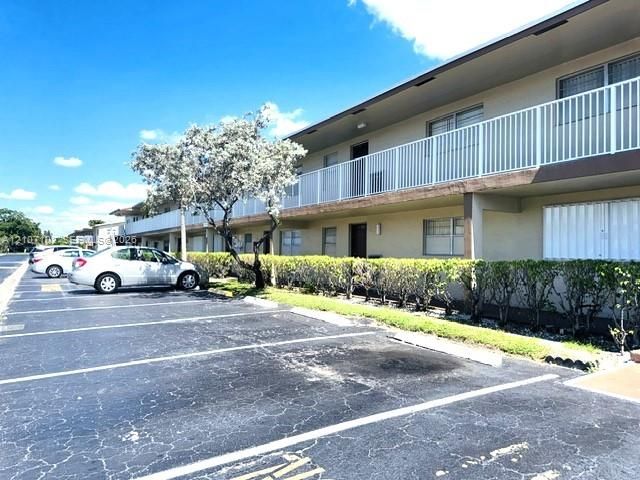 Multi-story exterior featuring a long balcony with white metal railings, light-colored stucco facade, horizontal window treatments, and asphalt surface parking