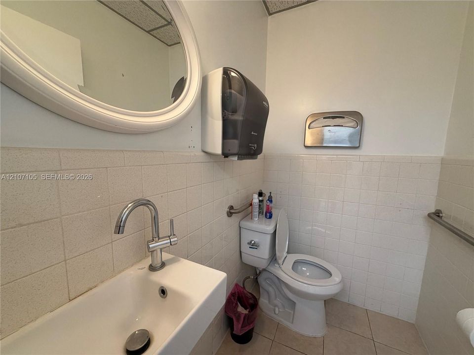 Wall-mounted sink with a chrome gooseneck faucet, oval mirror, square tile wainscoting, and stainless steel wall-mounted hardware