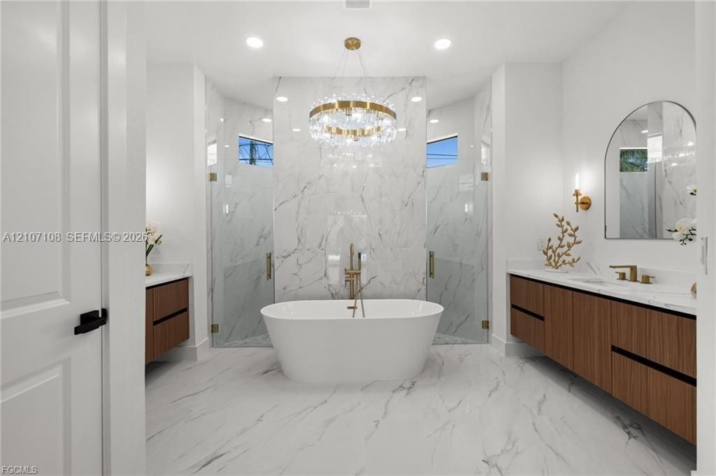 Freestanding white soaking tub, dual floating wood-finish vanities, wall-to-wall stone-surround shower, gold-tone plumbing fixtures, and a circular crystal chandelier