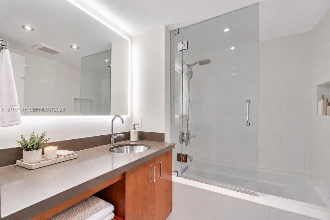 Bathroom featuring a wood-finish vanity with a stone countertop, an integrated sink, an illuminated mirror, and a glass-enclosed shower with a soaking tub