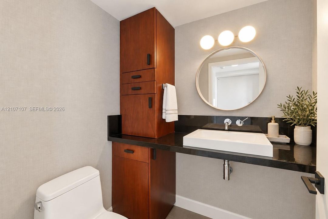 Built-in wood-finish cabinetry, black countertop, vessel sink, wall-mounted faucet, and circular vanity mirror with three-bulb integrated lighting