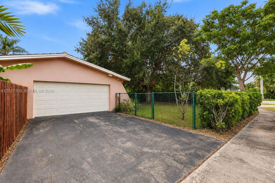 Attached garage with a wide sectional door, paved driveway, chain-link perimeter fencing, and mature trees on a grass-covered lot