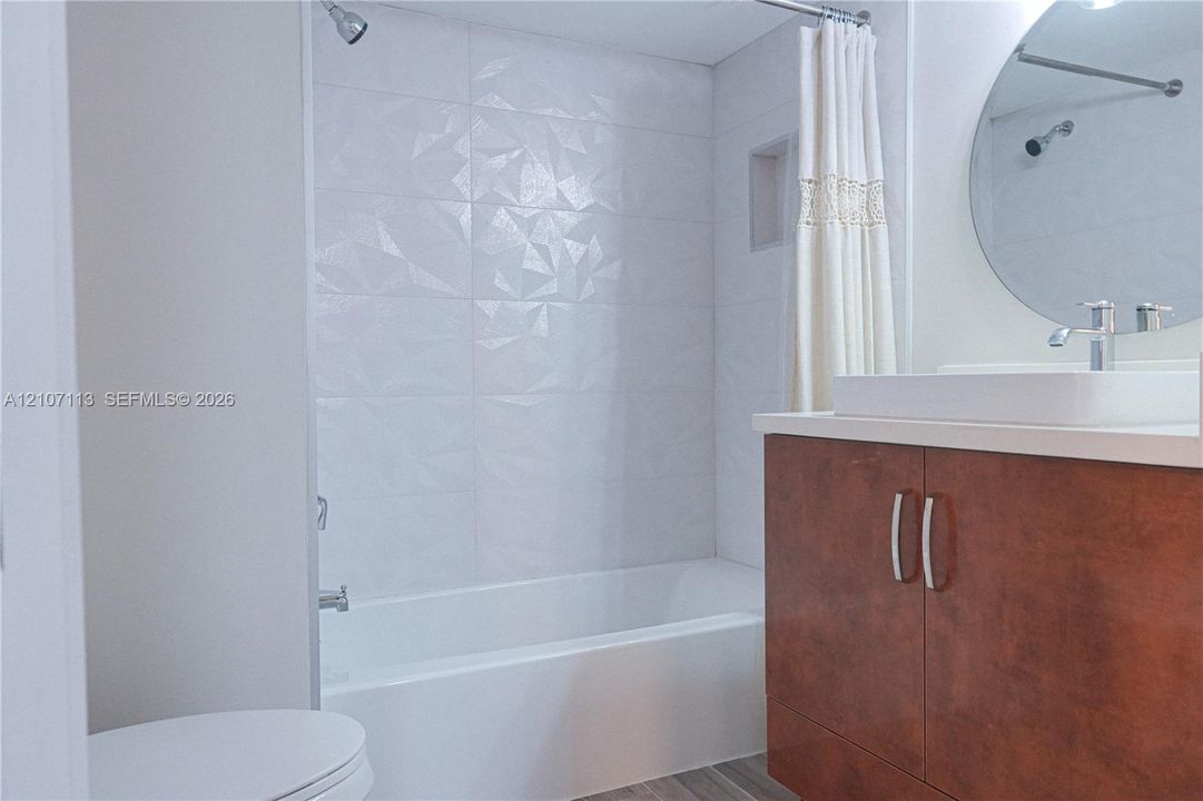 Bathroom featuring a wood-finish vanity with a rectangular basin, geometric textured tile shower surround, built-in shower niche, and a circular wall-mounted mirror