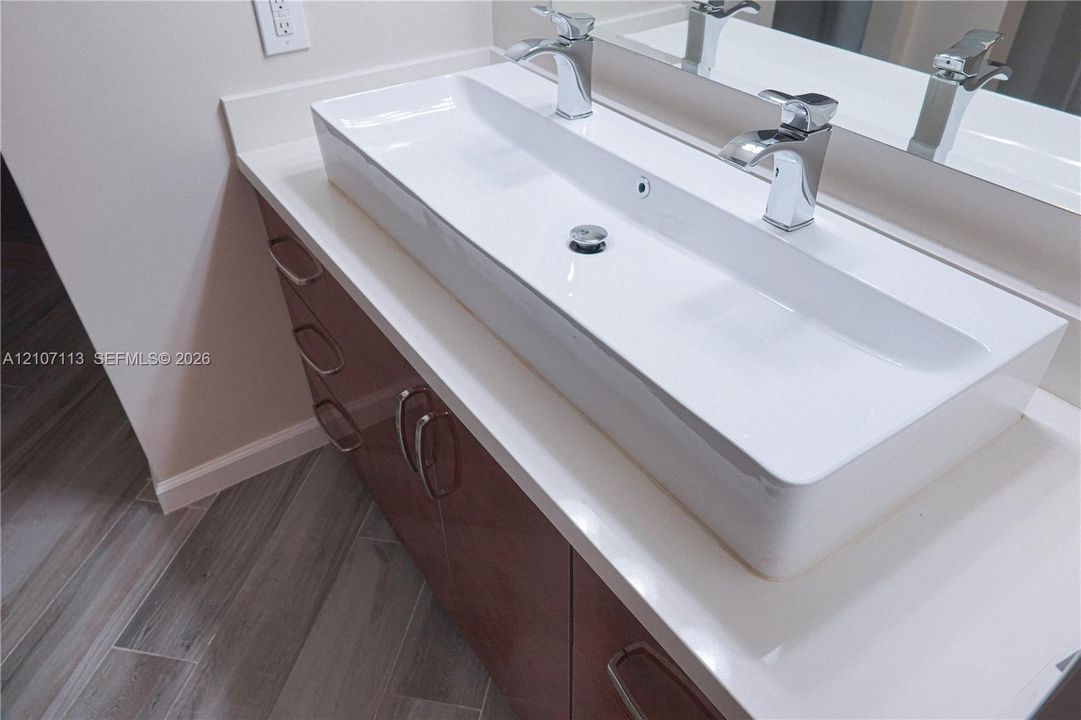 Bathroom vanity featuring a long, rectangular integrated sink, dual chrome faucets, solid-surface countertop, dark-toned cabinetry with metallic hardware, and wood-finish flooring
