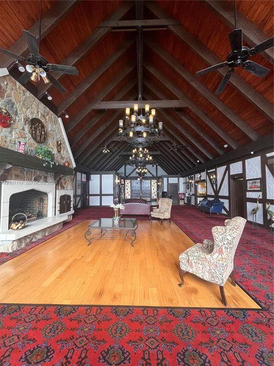 Vaulted ceiling with exposed wood beams, stone fireplace featuring an integrated hearth, central wood-finish flooring, wrought-iron chandeliers, and Tudor-style wall detailing