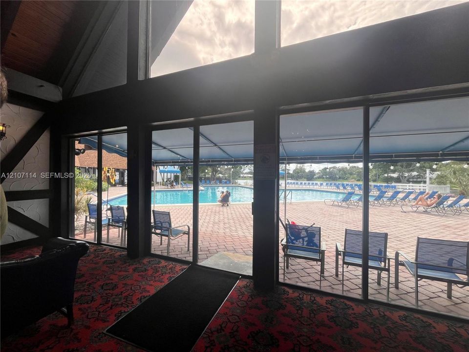 Floor-to-ceiling glass paneling, high-pitched timber-beamed ceiling, and direct access to an expansive paver-tiled pool deck featuring a covered outdoor awning