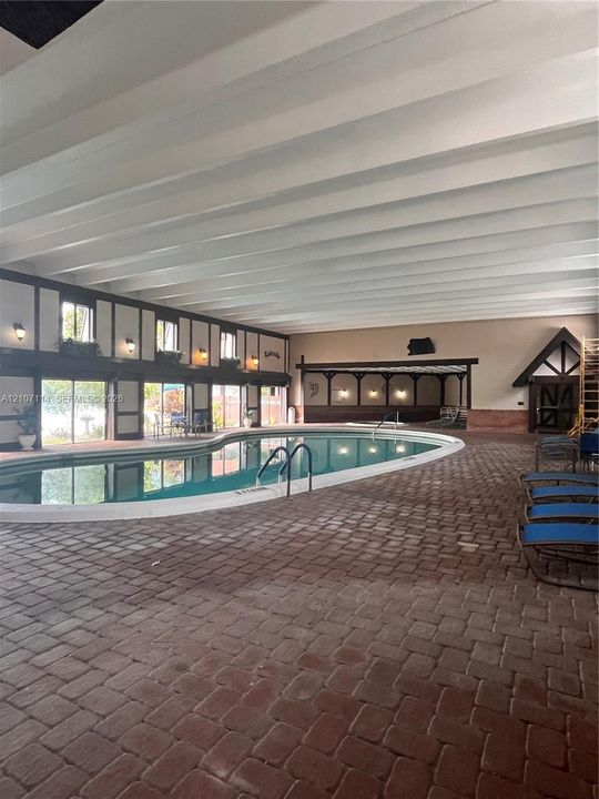 Indoor swimming area featuring a large curved pool, vaulted beamed ceiling, floor-to-ceiling windows, and brick paver flooring