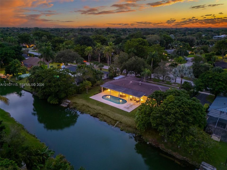 Waterfront property featuring an oval swimming pool, expansive paved patio, covered outdoor area, and mature landscape