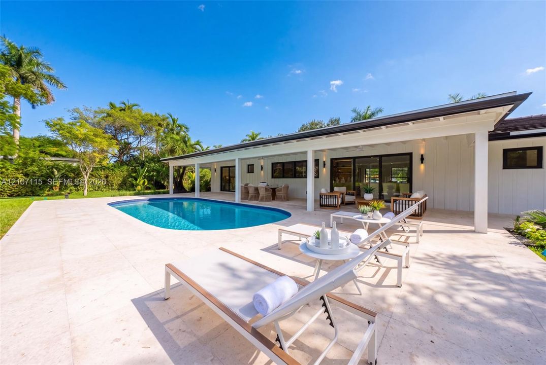 Outdoor living space featuring an expansive stone-tiled patio, a built-in swimming pool, a covered lanai, and white vertical board-and-batten siding