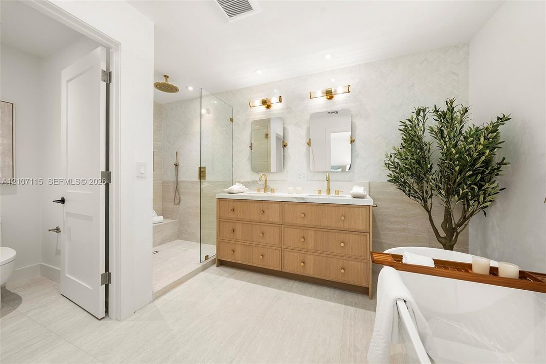 Double vanity with wood-finish cabinetry, countertop with integrated dual sinks, gold-tone wall-mounted lighting, glass-enclosed shower with tile surround, and freestanding soaking tub