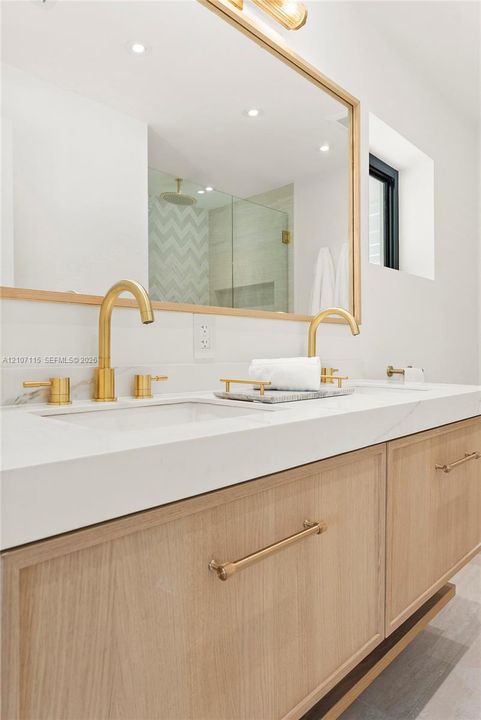 Floating vanity with light wood-finish cabinetry, solid surface countertop, dual undermount sinks, brass-toned hardware, and a framed mirror