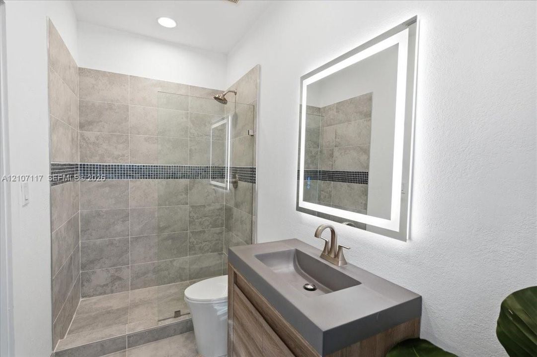 Bathroom featuring a wood-finish vanity with a molded sink, LED-backlit mirror, walk-in shower with large-format tile and a glass partition, and a mosaic tile inlay