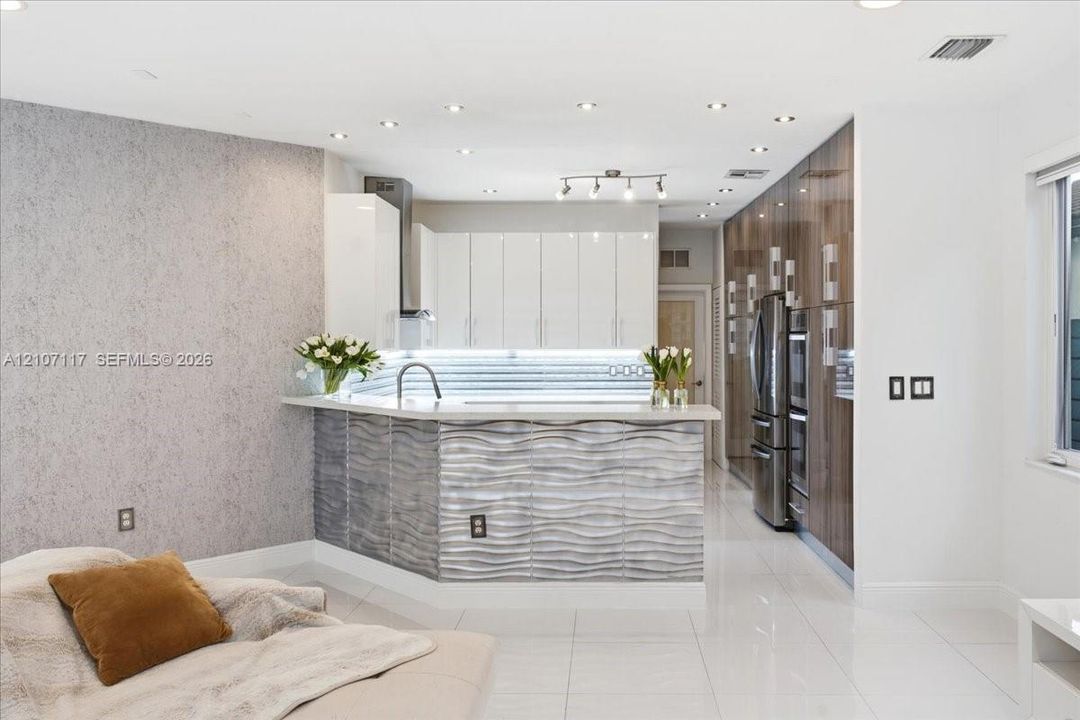 Contemporary kitchen featuring high-gloss white upper cabinetry, full-height wood-finish pantry storage, a raised counter with a textured wave-pattern facade, integrated stainless steel appliances, and polished tile flooring