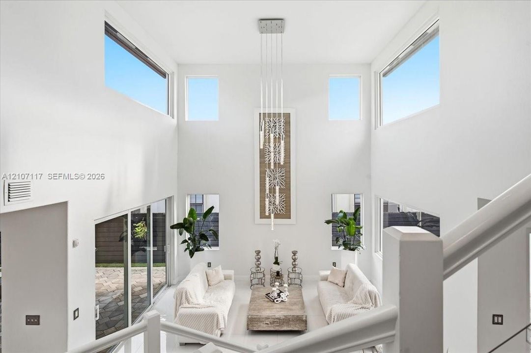 Double-height ceilings, expansive window configuration, multi-level glass slider access to exterior patio, and tiered contemporary pendant lighting