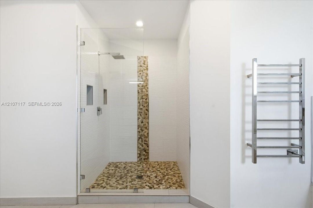 Walk-in shower featuring a frameless glass door, stone-tile flooring and wall accent, dual recessed wall niches, rainfall showerhead, and a wall-mounted metal towel warmer