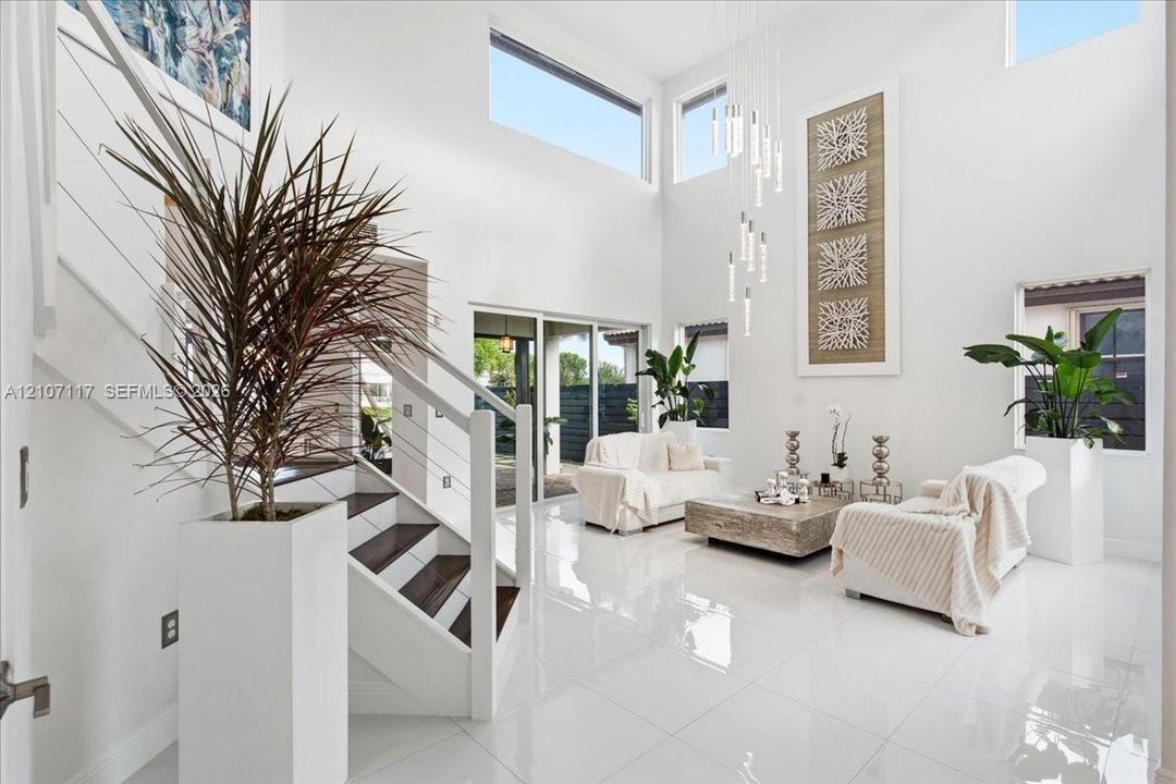 Double-height living area featuring floor-to-ceiling walls, polished oversized tile flooring, a multi-tiered glass pendant light fixture, and a staircase with a white-finished railing and wood-finish treads