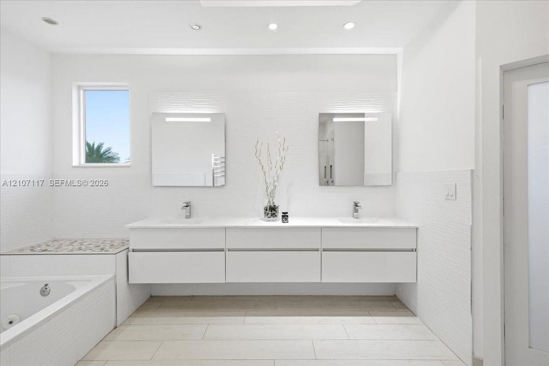 Floating white double vanity with polished stone countertop, rectangular undermount sinks, wall-mounted light fixtures, tiled bathtub surround with stone inlay, and tile flooring
