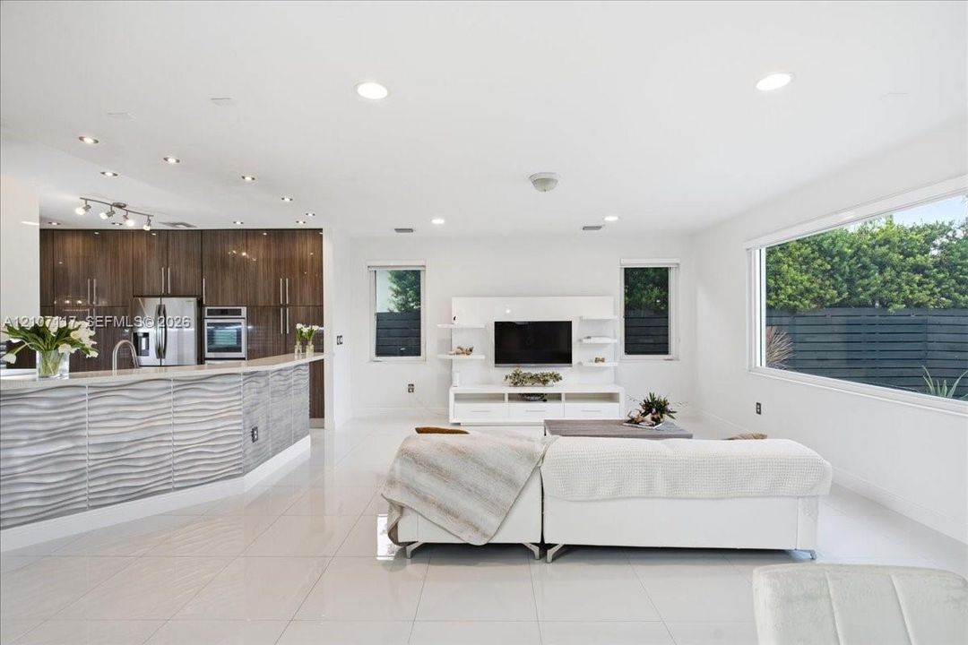 Open-concept interior featuring large-format tile flooring, recessed ceiling lighting, kitchen cabinetry with a horizontal wood-grain finish, and a peninsula with a textured, undulating gray facade