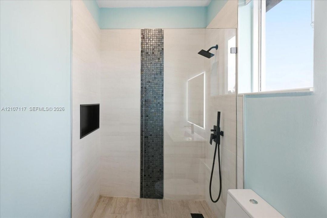 Walk-in shower featuring large-format wall tile, a vertical mosaic accent strip, matte black wall-mounted shower hardware, a recessed wall niche, and a glass partition
