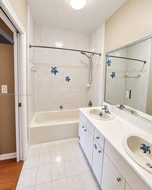 Dual-sink vanity with a solid-surface countertop and integrated basins, tile surround bathtub with a handheld shower fixture, and ceramic tile flooring