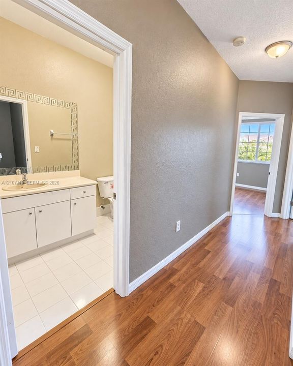 Hallway featuring wood-finish flooring and neutral-tone walls, with a bathroom containing white cabinetry, a single-basin vanity, and tile flooring