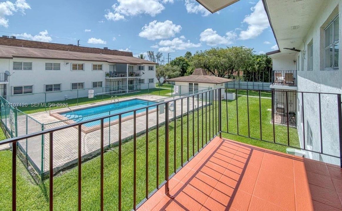 Balcony featuring tiled flooring and black metal railings, overlooking a fenced community swimming pool, concrete pool deck, and landscaped lawn