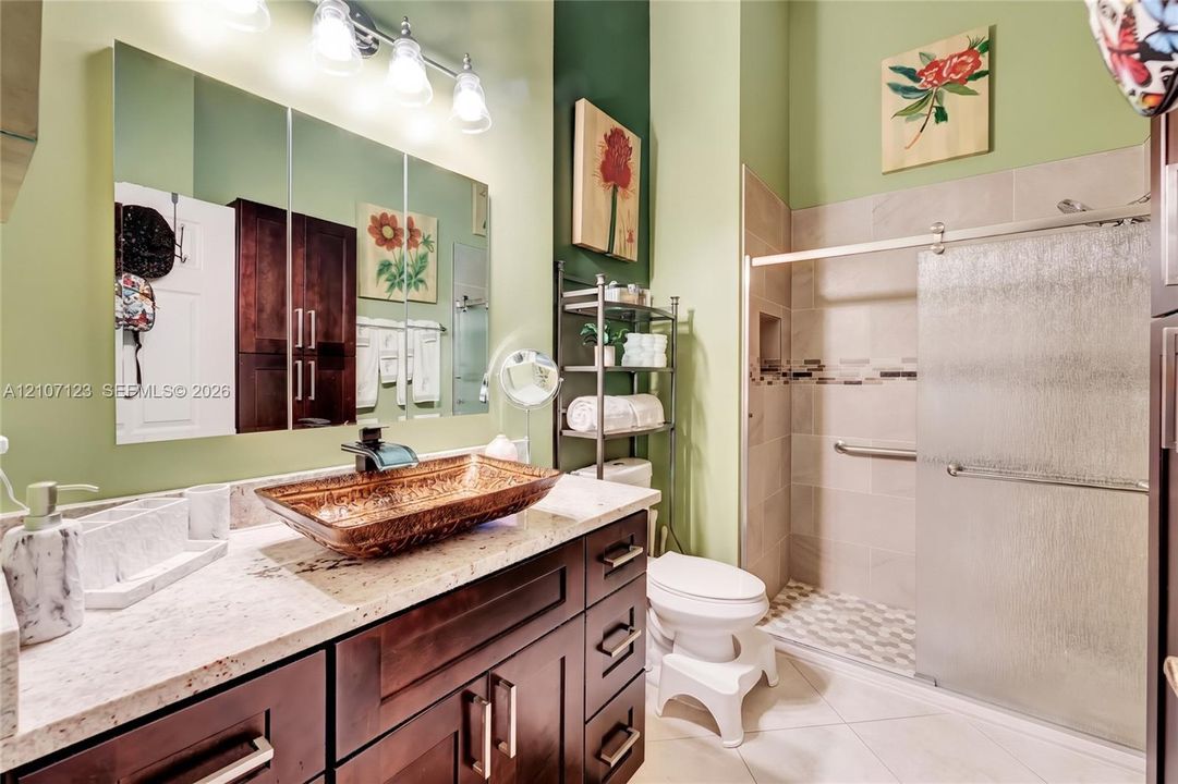 Bathroom featuring a dark wood vanity with stone-finish countertop, hammered metal vessel sink, four-light vanity fixture, and walk-in shower with sliding glass doors and wall-mounted grab bar