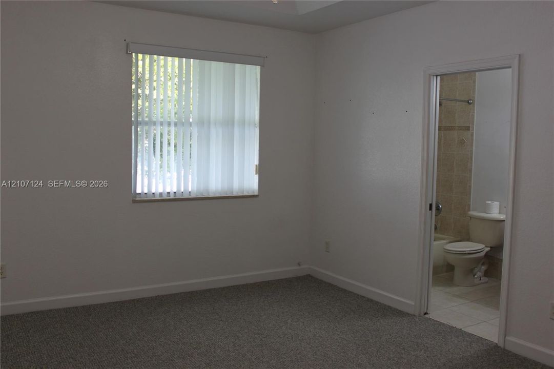 Room featuring neutral-toned carpeting, a window with vertical blinds, and a door frame opening to a bathroom with tile flooring and wall-mounted plumbing fixtures