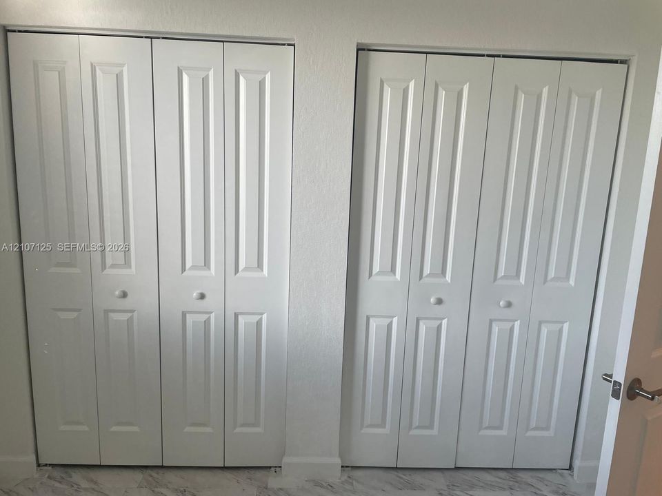 Dual sets of white bi-fold closet doors featuring recessed panel detailing and round knobs, complemented by neutral-toned stone-finish flooring