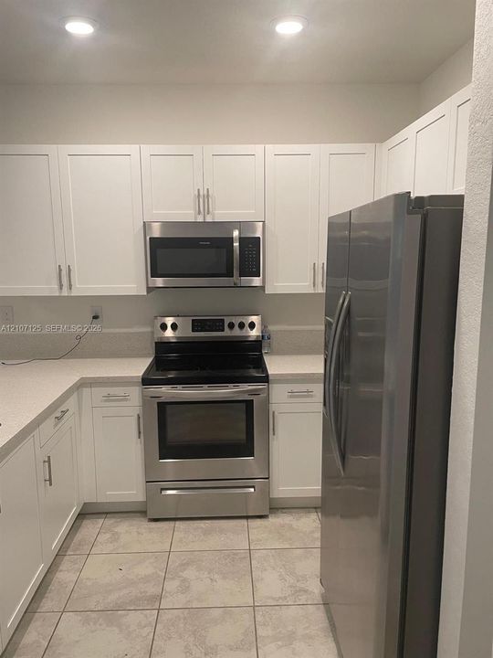 Kitchen featuring white shaker-style cabinetry, light-toned composite countertops, tile flooring, and stainless steel appliances including a built-in microwave, range, and refrigerator