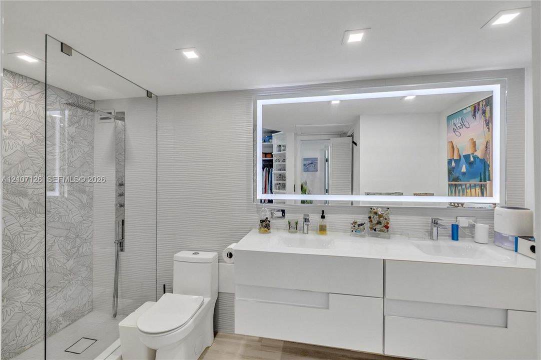 Floating dual-sink vanity with a solid-surface countertop, glass-enclosed walk-in shower with decorative tile detailing, large LED-backlit mirror, and recessed ceiling lighting