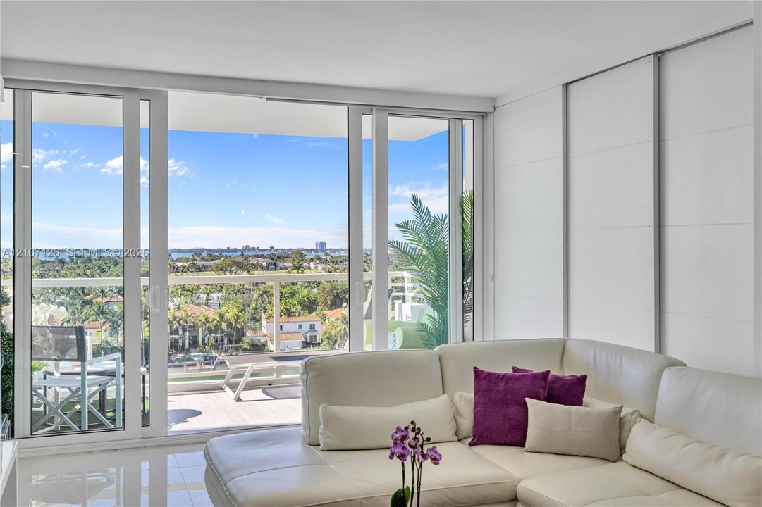 Floor-to-ceiling sliding glass doors opening to a covered balcony with a glass railing, white polished flooring, and expansive exterior water and landscape scenery