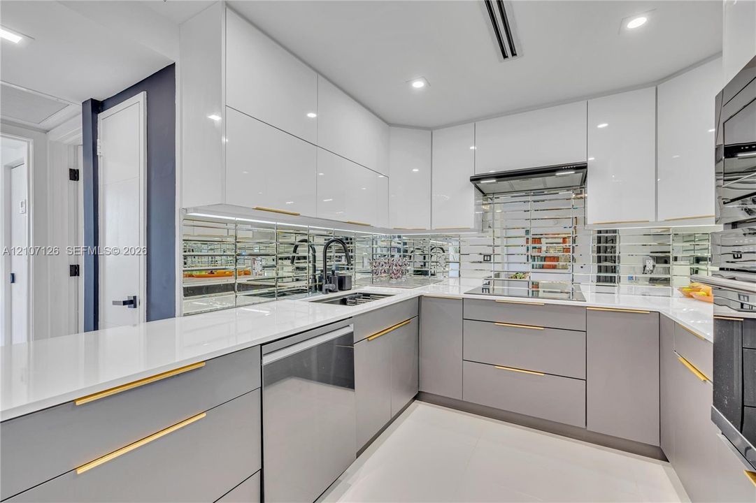 Modern kitchen featuring high-gloss white upper cabinetry, grey flat-panel lower cabinetry, gold-tone hardware, white stone countertops, and a mirrored tile backsplash