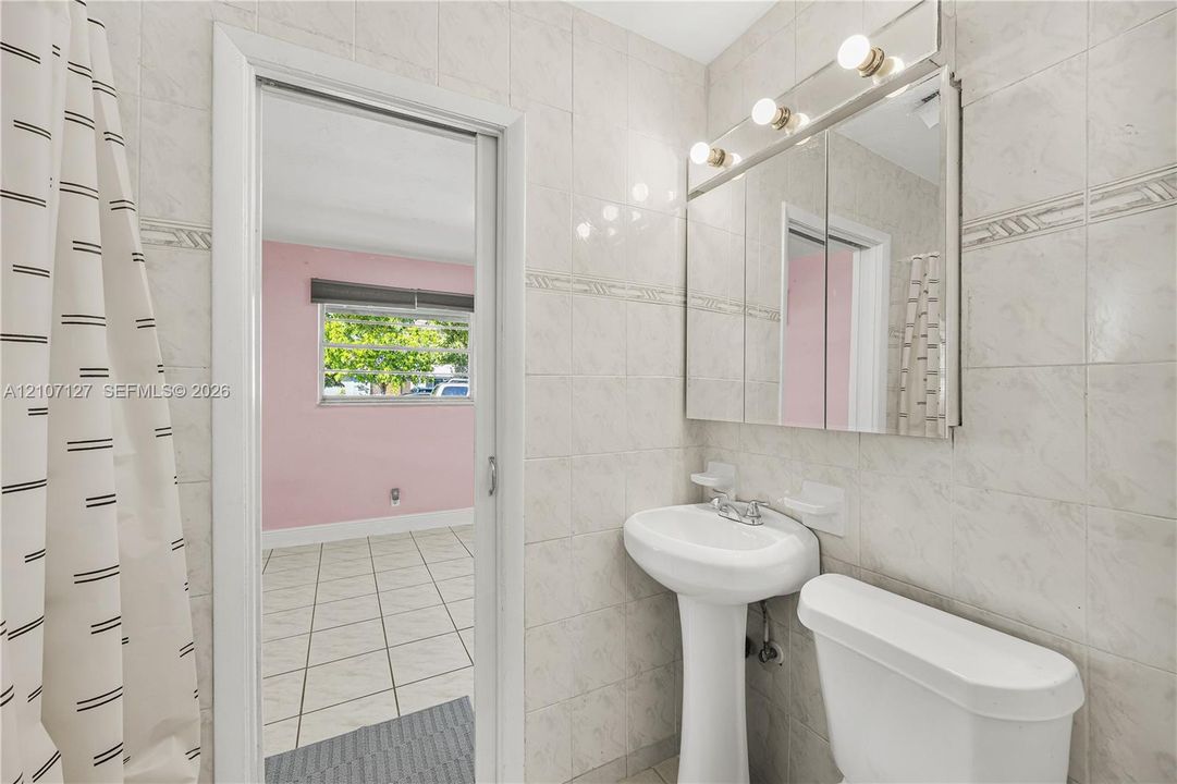 Full-height tile wall surround, white porcelain pedestal sink, mirrored medicine cabinet with integrated vanity lighting, and standard toilet