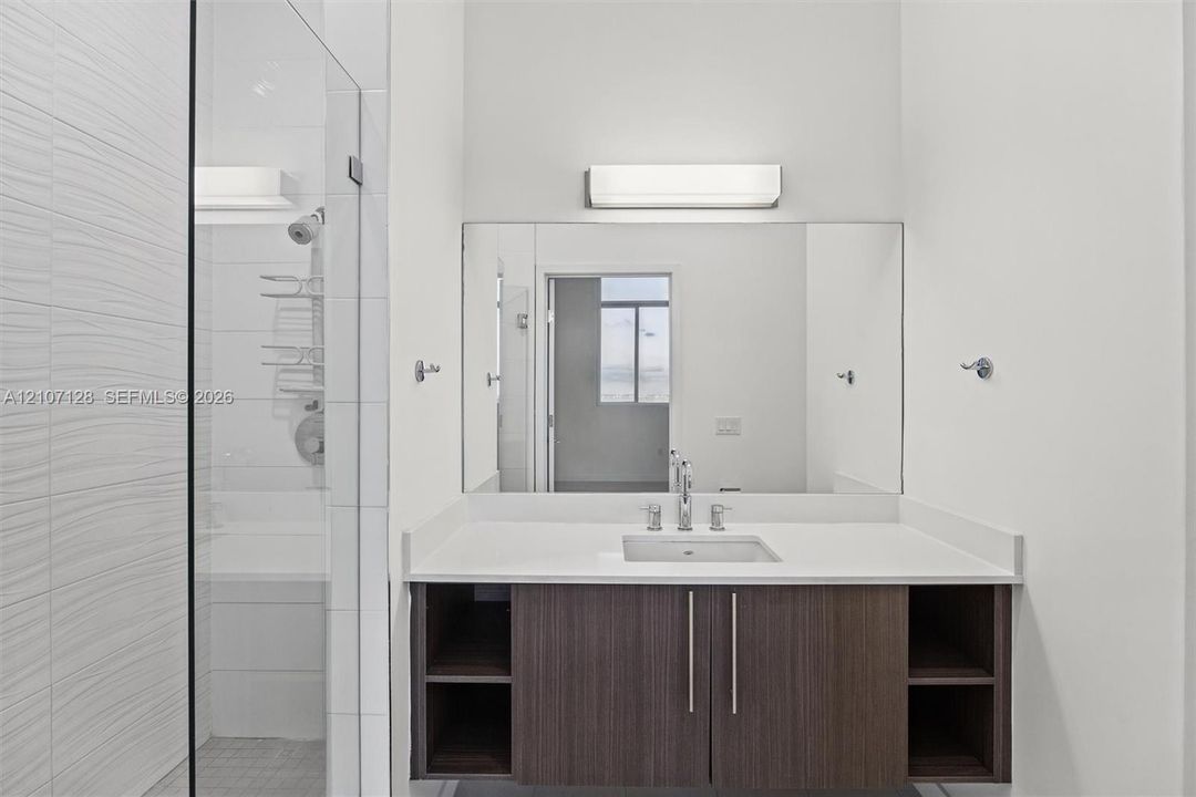Private bathroom with wall-mounted vanity, wood-finish cabinetry with open side shelving, white countertop with an integrated rectangular sink, a polished chrome faucet, a large frameless mirror, and a glass-enclosed shower with textured wall tile