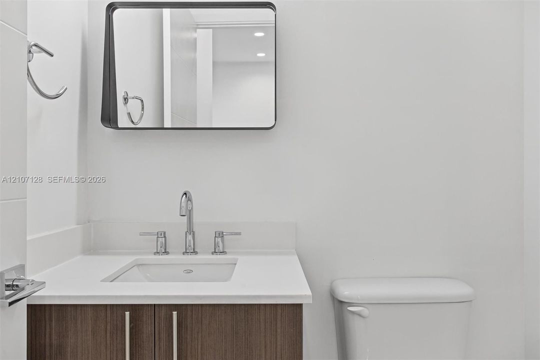 Powder room with wood-finish cabinetry, stone-surface countertop, rectangular undermount sink, chrome widespread faucet, and a wall-mounted mirror