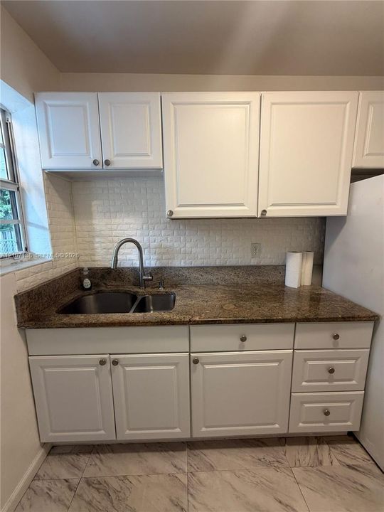 Kitchen featuring white shaker-style cabinetry, stone-finish countertops, dual-basin stainless steel sink with gooseneck faucet, textured tile backsplash, and light-tone tile flooring