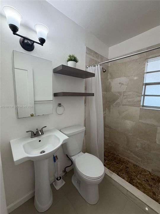 Bathroom featuring a pedestal sink with two-handle faucet, wall-mounted light fixture, floating shelving, and a shower enclosure with floor-to-ceiling neutral tile surround