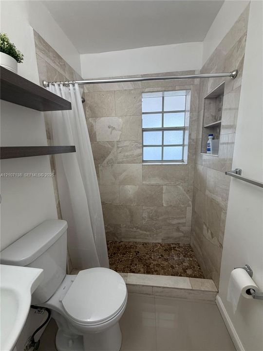 Walk-in shower featuring stone-tiled walls, a pebble-stone floor, a built-in recessed shelf, and a glass-block window