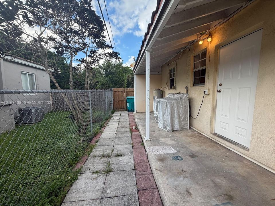 Covered concrete patio featuring a tiled walkway, chain-link perimeter fencing, and exterior wall-mounted lighting