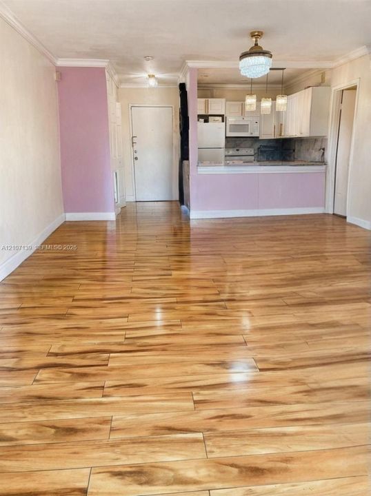 Open-concept area featuring wood-finish flooring, crown molding, tiered pendant lighting, and a kitchen pass-through with white cabinetry
