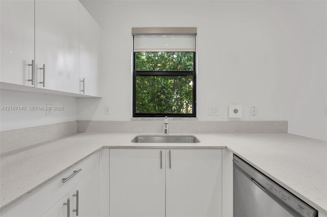 Kitchen featuring white flat-panel cabinetry, light-toned composite countertops with matching backsplash, undermount stainless steel sink with pull-down faucet, and integrated stainless steel dishwasher