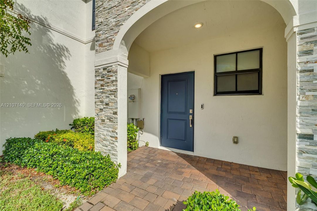 Covered entryway featuring a stone veneer accent pillar, a dark blue solid-panel door, and paved brick flooring